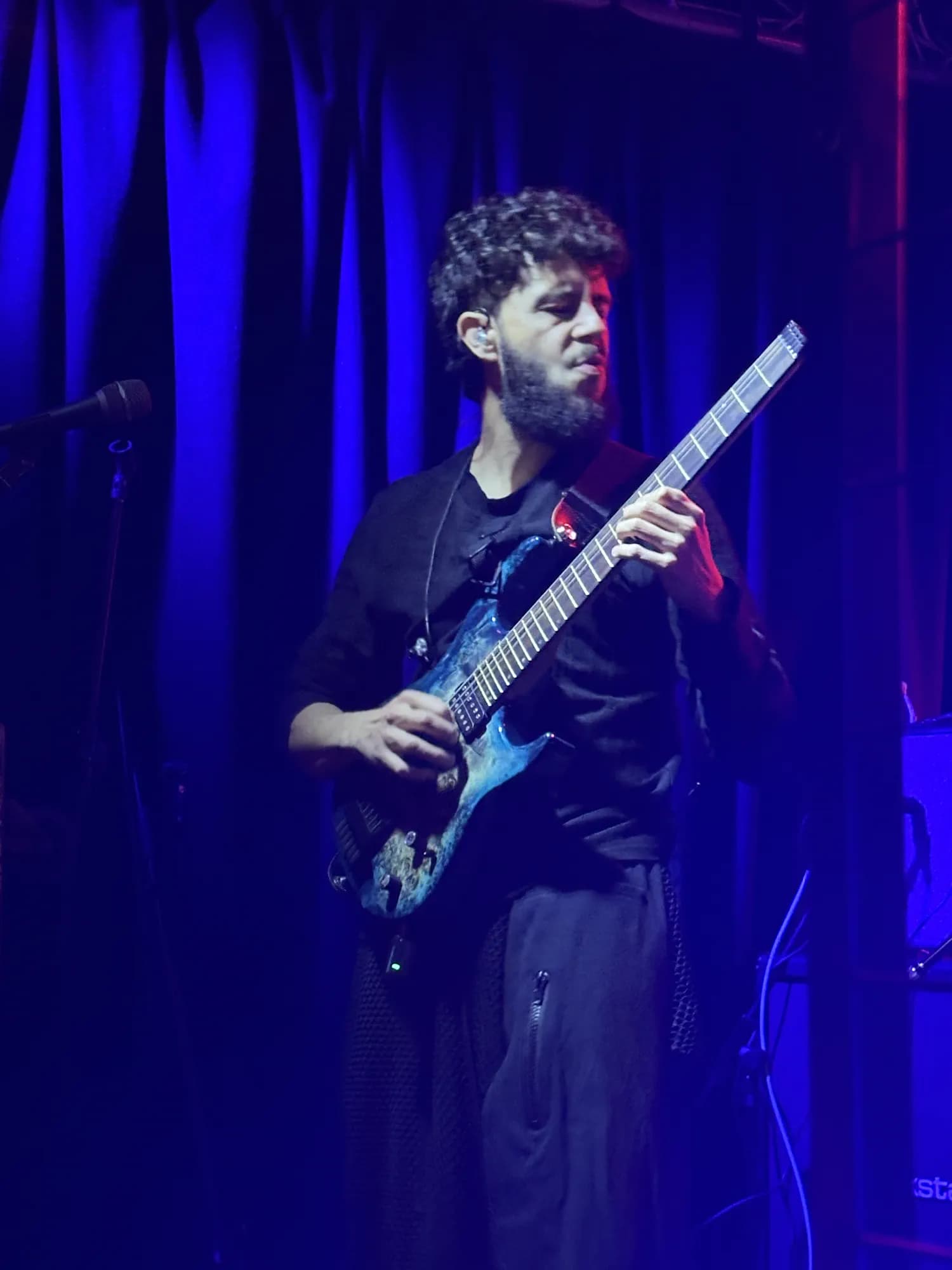 The guitarist mid-riff on a blue-lit stage, working the neck of a burl-topped electric guitar, a mic stand beside him.