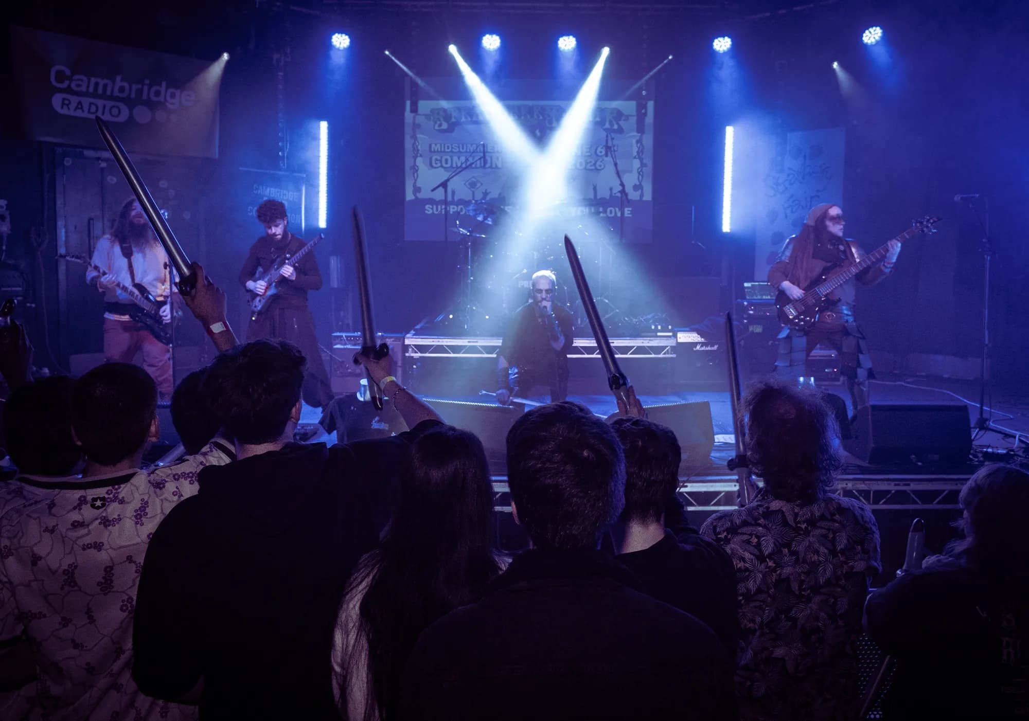 From the floor, the crowd lifts foam swords toward the band as they play under sweeping blue light.