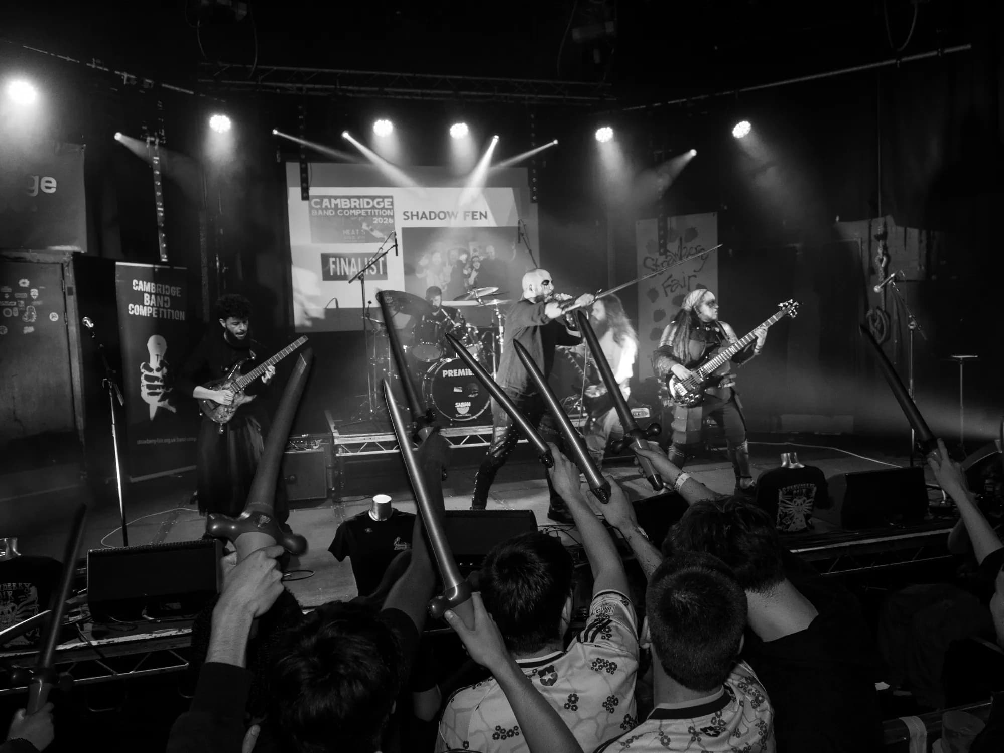 Shadow Fen command the stage in full cry as the crowd raises foam swords, the Cambridge Band Competition screen glowing behind them.