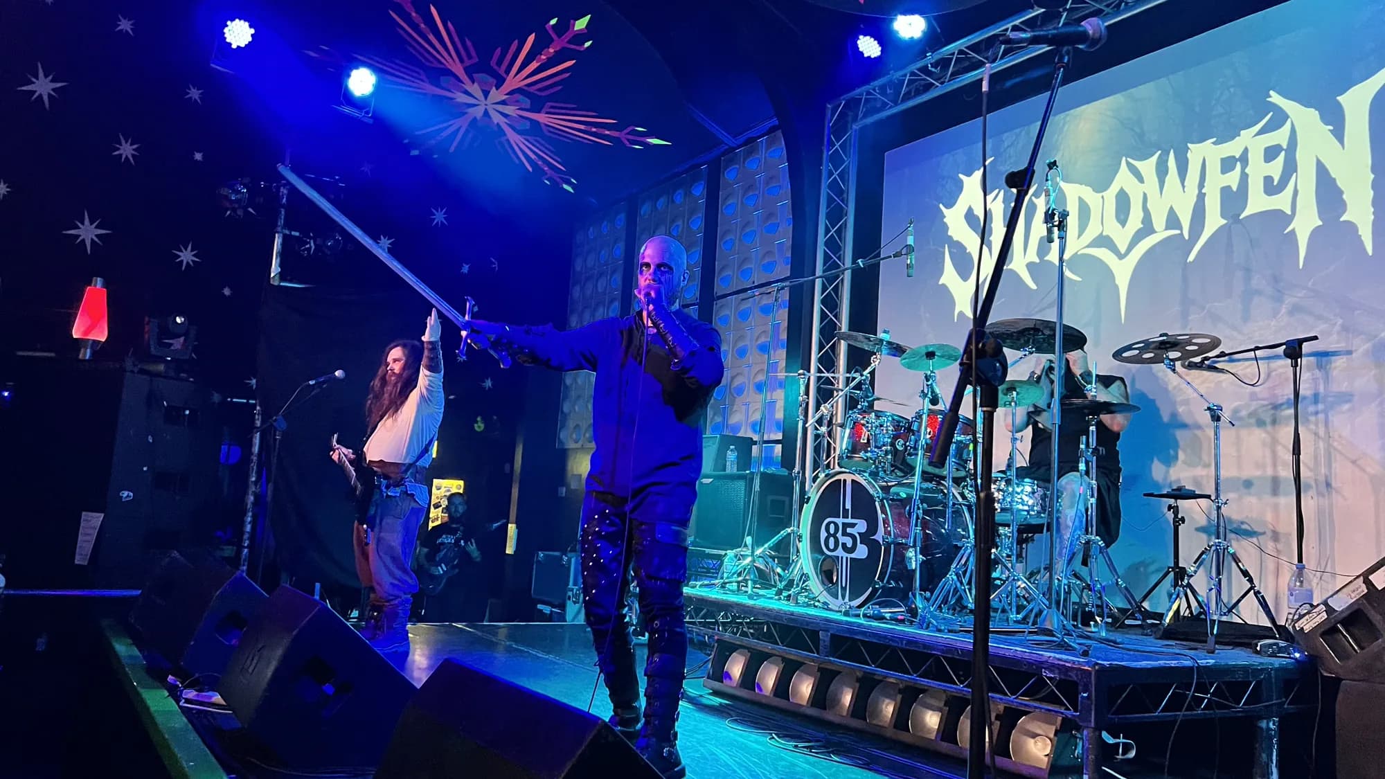 The full band on a blue-lit stage beneath their Shadow Fen backdrop, the vocalist raising a sword above his head.